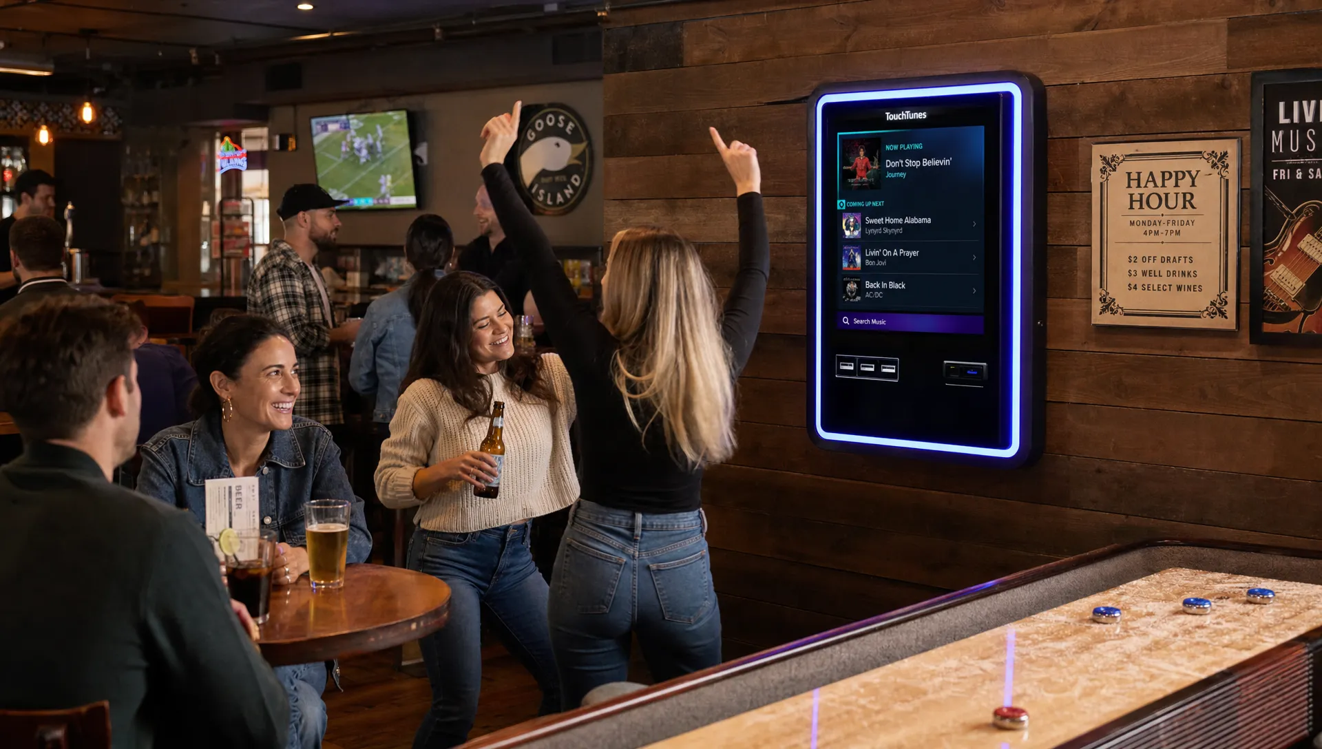 A medium interior view of a bar with guests at tables, a wall-mounted digital jukebox, and people reacting to music while drinks and a bar game table sit nearby in a social atmosphere.